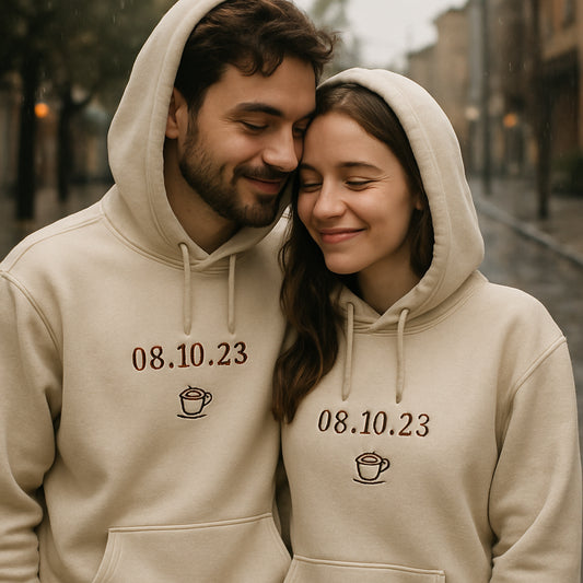 A cozy couple wearing matching custom hoodies on a rainy street, showing detailed embroidery of a date and tiny coffee cup icon. Alt: Trendy design ideas for custom couple hoodies
